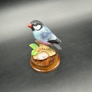 Könnte beinhalten: Eine Keramik-Vogelfigur mit grauem Kopf, hellblauem Körper und schwarzen Flügeln. Der Vogel sitzt auf einem Ast mit grünen Blättern und orangen Beeren, montiert auf einem Holzsockel. Der Schnabel des Vogels ist rot.