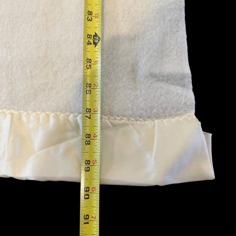 May include: A white blanket with a satin border. The blanket is measured with a yellow measuring tape. The blanket is approximately 83 to 91 inches long.
