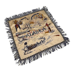 May include: Cream-colored square throw blanket with a fringed border. The blanket features a vintage illustration of Alaska, including the text "Alaska Land of the Midnight Sun" and various Alaskan imagery.