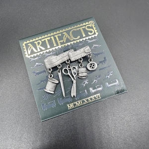 May include: A silver-toned brooch featuring miniature sewing tools, including scissors, a needle, a spool of thread, and a thimble, suspended from a ruler design. The brooch is set against a dark green background with the word "ARTIFACTS" and Roman numerals.