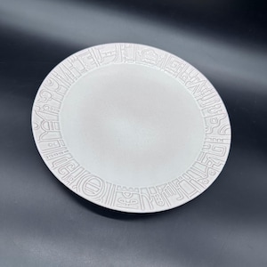 May include: A white ceramic plate with a raised, repeating geometric pattern around the rim. The plate has a smooth, slightly textured surface and a simple, elegant design. The plate is suitable for serving food.