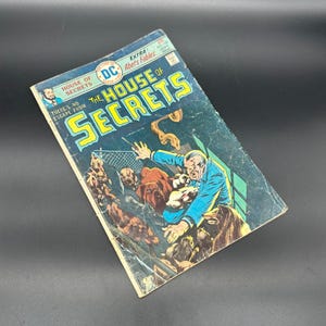 May include: A vintage comic book cover featuring a man in a blue suit fighting off a group of monstrous creatures. The cover is titled "The House of Secrets" and features the text "There's no escape from the House of Secrets".