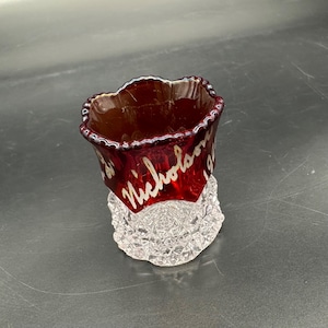 Puede incluir: Una pequeña copa de vidrio rojo con una base de vidrio transparente. La copa tiene un borde festoneado y está decorada con letras doradas que dicen "St. Nicholas 192".