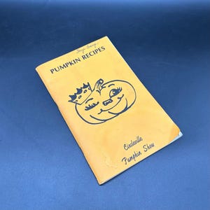 May include: A vintage cookbook titled "Pumpkin Recipes" with a smiling pumpkin illustration on the cover. The cover is orange with black text and a black pumpkin face wearing a crown. The book is slightly worn.