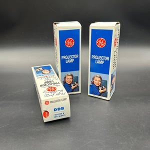 May include: Three vintage GE Projector Lamp boxes. The boxes are white with blue panels and feature a woman holding a projector lamp. The boxes have the text "PROJECTOR LAMP" and "DDB 115-120 V 750 WATTS".
