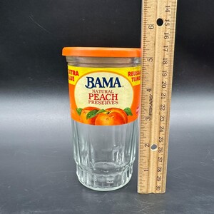 May include: A clear glass jar with an orange lid and a label that reads "BAMA NATURAL PEACH PRESERVES." The jar is approximately 5 inches tall and has vertical ridges. A wooden ruler is next to the jar.