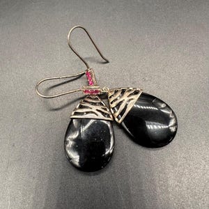 May include: A pair of drop earrings featuring smooth, teardrop-shaped black stones suspended from silver-toned metal caps with a textured, open-work design. Each earring is accented with small, vibrant pink stones set into the metal hook attachment.