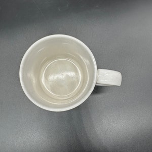 May include: A white ceramic mug with a handle. The mug is empty and has a smooth, glossy finish.