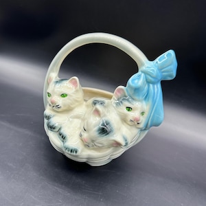 May include: A ceramic basket-shaped planter featuring three white cats with gray markings and green eyes. The basket has a handle and a light blue bow on the side. The planter is a decorative item.