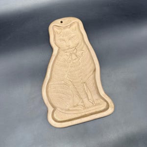 May include: A beige, cat-shaped cookie mold with a detailed embossed design. The cat is seated, wearing a bow and bells. The mold has a hole at the top for hanging. The overall design is a vintage style.