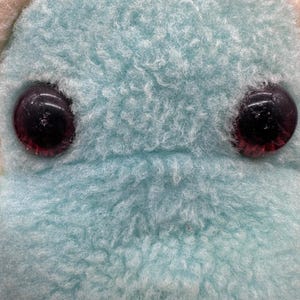 Peut inclure: Gros plan sur le visage d'une peluche. Le jouet a deux grands yeux ronds et sombres avec des reflets rouges. La fourrure est d'un bleu clair et doux. La texture de la fourrure est duveteuse et l&eacute;g&egrave;rement feutr&eacute;e.