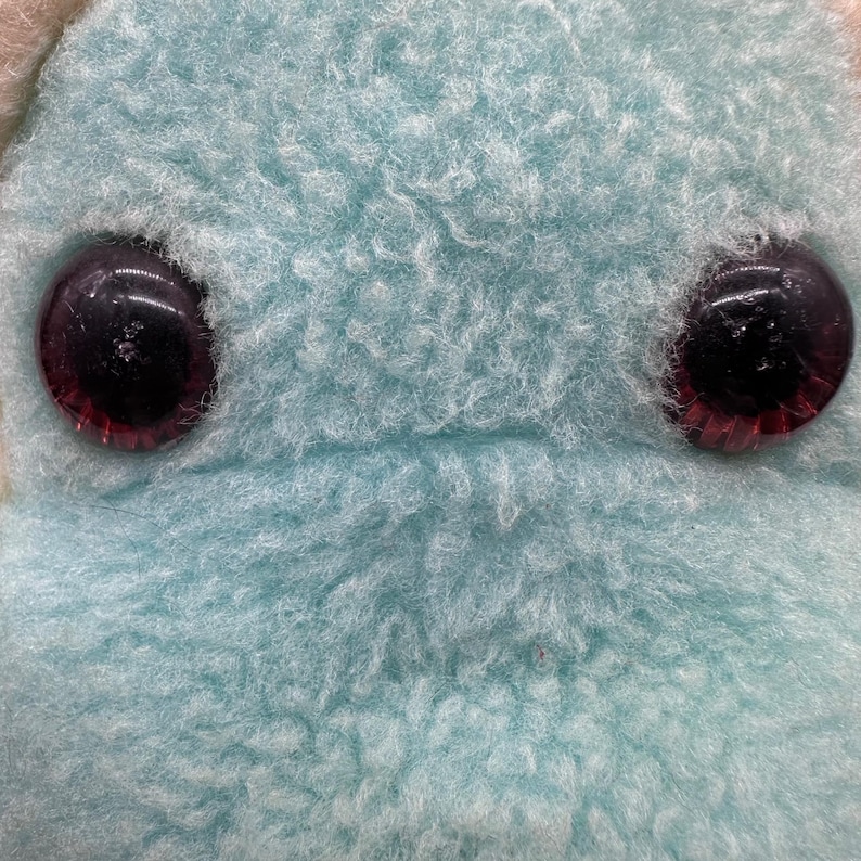 Peut inclure: Gros plan sur le visage d'une peluche. Le jouet a deux grands yeux ronds et sombres avec des reflets rouges. La fourrure est d'un bleu clair et doux. La texture de la fourrure est duveteuse et l&eacute;g&egrave;rement feutr&eacute;e.