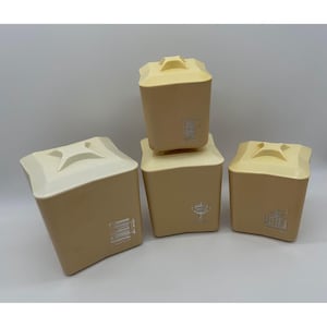 Puede incluir: Cuatro botes cuadrados beige y crema con tapas. Los botes tienen una superficie texturizada y gráficos decorativos plateados. Las tapas son de un color crema más claro y tienen un asa. Los botes son de diferentes tamaños.