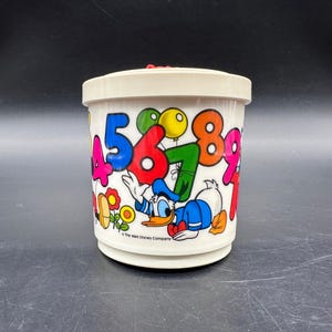 Puede incluir: Un recipiente de pl&aacute;stico blanco vintage con tapa, con n&uacute;meros coloridos y una imagen de dibujos animados de Donald Duck. Los n&uacute;meros son de varios colores, y el recipiente tiene una base y una tapa. The Walt Disney Company est&aacute; impreso.