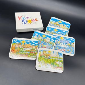 Puede incluir: Un juego de seis posavasos cuadrados con ilustraciones de paisajes urbanos y una caja a juego. Los posavasos presentan dibujos coloridos de varias ciudades, incluyendo Sídney y Melbourne, con cielos azules y soles amarillos. La caja dice "Ken Done".