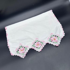 以下が含まれることがあります： 刺繍されたピンクのバラと緑の葉が特徴の、スカラップエッジの白い長方形の枕カバー。エッジはピンクのかぎ針編みでトリミングされています。枕カバーは柔らかい白い生地で作られています。