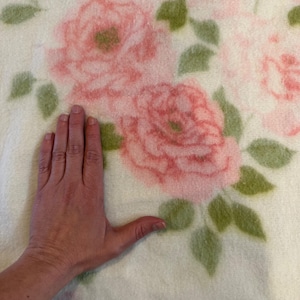 May include: A cream-colored blanket featuring a large floral pattern of pink roses and green leaves. The blanket's soft texture is visible, with a hand placed on the blanket for scale. The blanket is likely made of a soft material.