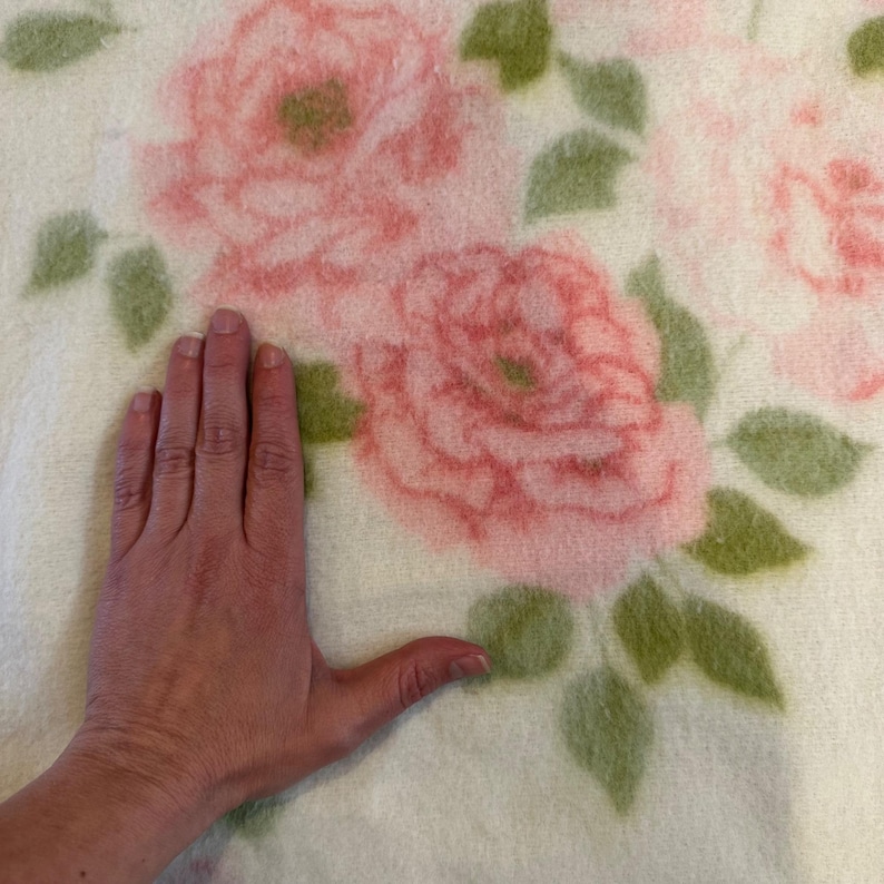 May include: A cream-colored blanket featuring a large floral pattern of pink roses and green leaves. The blanket's soft texture is visible, with a hand placed on the blanket for scale. The blanket is likely made of a soft material.