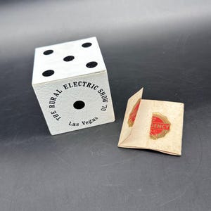 May include: A white die with black dots and the text "THE RURAL ELECTRIC SHOW '70 Las Vegas". Beside it is a small folded paper with a red and gold seal that reads "EMERGENCY".
