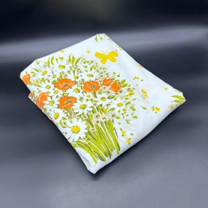 Puede incluir: Tela blanca con un diseño floral con amapolas naranjas, margaritas blancas y tallos verdes. También hay una mariposa amarilla. La tela está doblada, mostrando el patrón sobre un fondo oscuro.