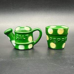 Puede incluir: Saleros y pimenteros de cerámica verde. El salero tiene forma de regadera con la palabra "SALT" escrita. El pimentero es una maceta con la palabra "PEPPER" escrita. Ambos tienen lunares blancos.