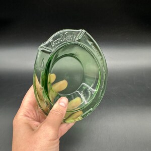 May include: A vintage green glass ashtray with the words "Good Luck" embossed on the rim. The ashtray has a circular center and a curved, horseshoe-like shape. The glass has a slight transparency.