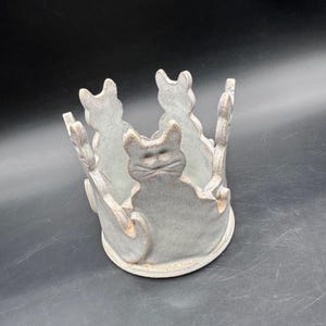 May include: A ceramic candle holder with a light gray finish. The holder features a circular design with cat silhouettes around the perimeter. The cats are in a sitting position, and the holder has a rustic, handmade appearance.