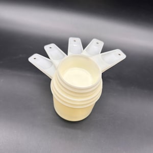 Puede incluir: Un juego de cinco tazas medidoras de plástico color blanco hueso apiladas. Cada taza cuenta con un recipiente redondo y un mango plano con un pequeño orificio circular en el extremo para colgar. Las tazas están encajadas y tienen bordes lisos.