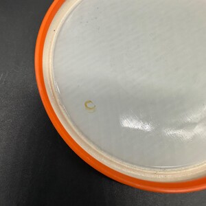 May include: A white disc golf disc with an orange rim. The disc has a smooth surface with a few minor imperfections. The orange rim is a bright, solid color. The disc is designed for recreational use.