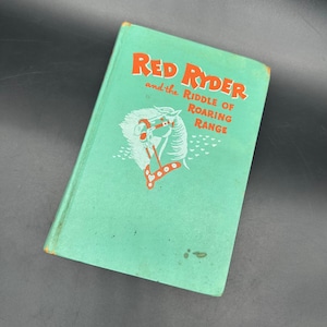 May include: A vintage book titled "Red Ryder and the Riddle of Roaring Range." The cover is a faded teal colour with orange text and a white illustration of a horse's head. The book shows signs of wear.