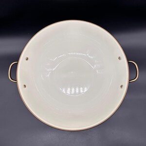 Op de afbeelding: Een ronde kookpan met een wijde opening, een gladde, cr&egrave;mekleurige binnenkant en een metalen, koperkleurige buitenrand. De pan is voorzien van twee gebogen, metalen handgrepen die met zichtbare klinknagels aan de zijkanten zijn bevestigd.