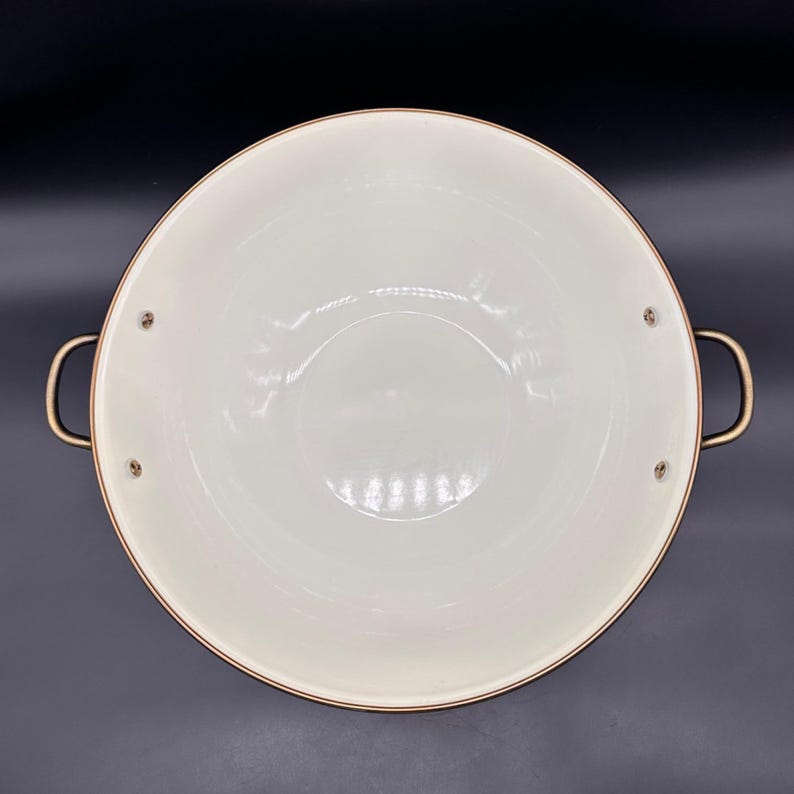 Op de afbeelding: Een ronde kookpan met een wijde opening, een gladde, cr&egrave;mekleurige binnenkant en een metalen, koperkleurige buitenrand. De pan is voorzien van twee gebogen, metalen handgrepen die met zichtbare klinknagels aan de zijkanten zijn bevestigd.