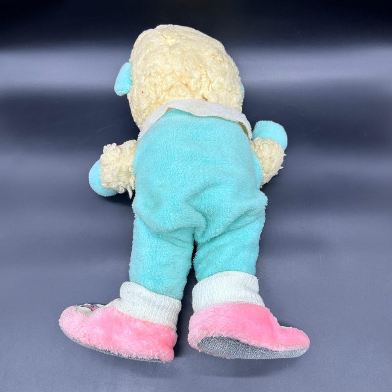 Peut inclure: Poup&eacute;e en peluche vintage, vue de dos, avec une t&ecirc;te et des membres jaune clair, et une combinaison turquoise. La poup&eacute;e porte des chaussettes blanches et des chaussons roses avec des motifs floraux. La fourrure de la poup&eacute;e est douce et textur&eacute;e.