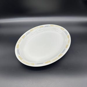 May include: White oval serving platter with a gold and black geometric border design. The dish is made of ceramic and is suitable for serving food. The platter is a classic piece for any dining table.