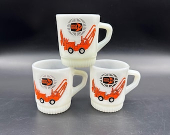 May include: Three white ceramic mugs stacked on top of each other. Each mug features an orange construction vehicle graphic and the words "Ditch Witch" inside a globe. The mugs have a handle and a ridged base.