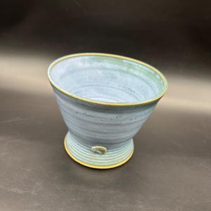 May include: A handmade ceramic bowl with a light blue glaze and a yellow rim. The bowl has a unique shape, with a flared top and a pedestal base. The bowl is approximately 5 inches in diameter and 4 inches tall.