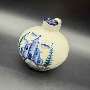 Puede incluir: Una jarra de cerámica redonda de color blanco roto con una pequeña asa y un cuello estrecho bordeado en azul oscuro. El cuerpo presenta una escena pintada a mano en azul y bronceado que representa una estructura tipo pagoda y pinos verdes.
