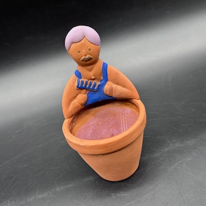 May include: A terracotta-colored ceramic planter with a figure of a person wearing blue overalls and a lavender hairstyle, holding a comb. The inside of the planter is a deep purple color.