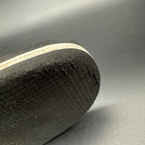 May include: Close-up of the sole of a black shoe with a white stripe. The sole has a textured pattern and shows signs of wear and tear, with some areas of the material peeling away. The shoe's design is simple and functional.