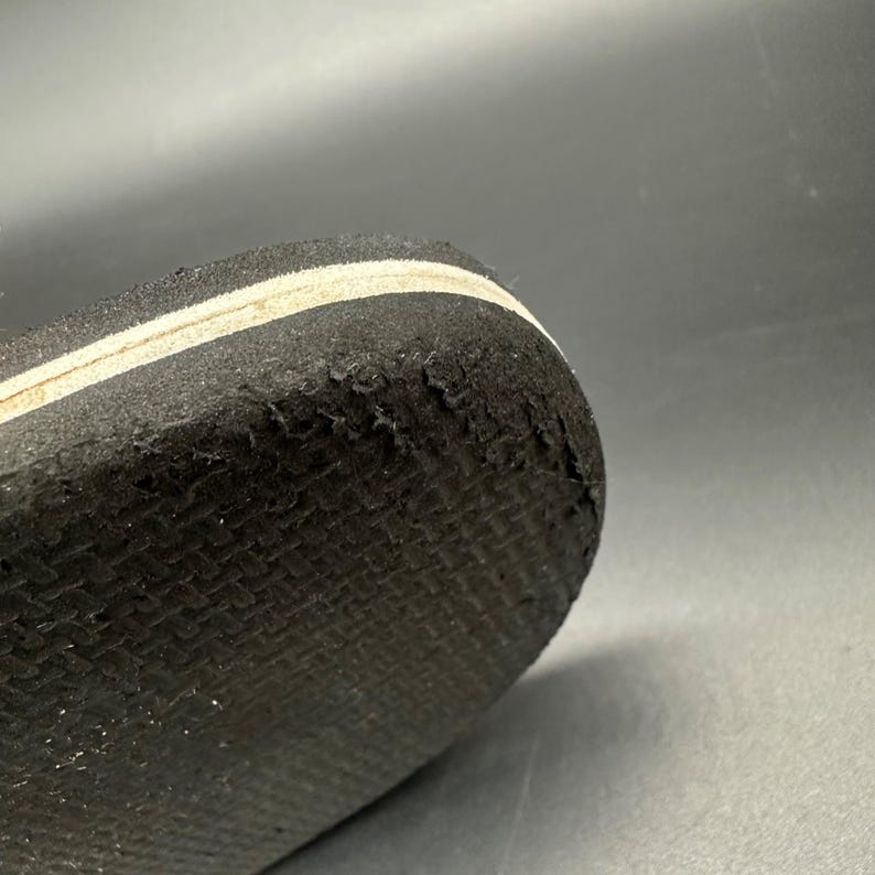 May include: Close-up of the sole of a black shoe with a white stripe. The sole has a textured pattern and shows signs of wear and tear, with some areas of the material peeling away. The shoe's design is simple and functional.