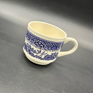 May include: A white ceramic mug with a blue and white willow pattern. The mug has a handle and a wide rim.