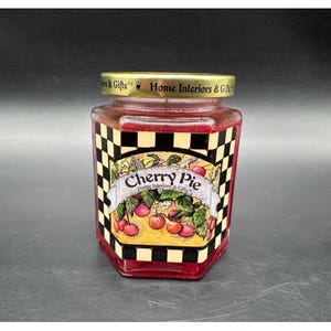 Peut inclure: Un pot en verre hexagonal rempli d'une substance rouge, peut-être une bougie ou de la gelée. Le pot a un couvercle doré et une étiquette qui dit "Cherry Pie" avec une illustration de cerise. L'étiquette est encadrée d'un motif à carreaux noir et blanc.