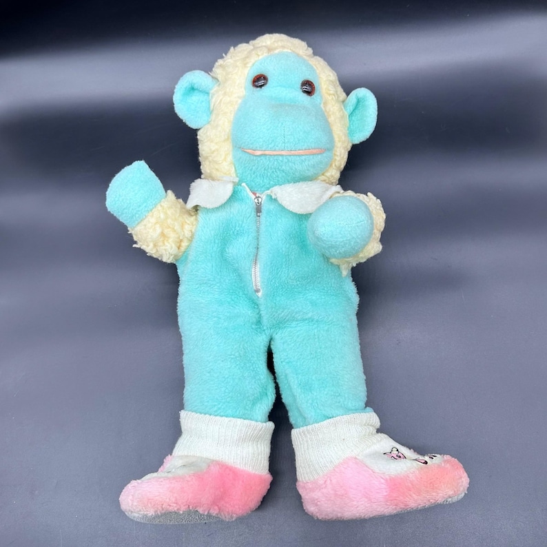 Peut inclure: Un singe en peluche vintage, v&ecirc;tu d'une combinaison bleu clair avec une fermeture &eacute;clair, un col blanc et des moufles assorties. Le singe a une t&ecirc;te et des oreilles jaune clair, avec des traits bleus. Les pieds sont blancs avec des semelles roses et des d&eacute;tails brod&eacute;s.