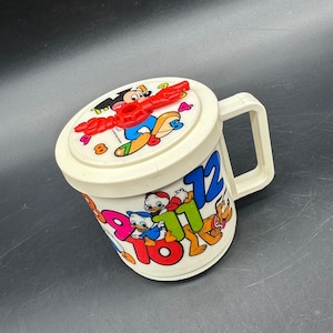 Puede incluir: Una taza de pl&aacute;stico crema vintage con tapa que presenta un dise&ntilde;o de reloj de Mickey Mouse. La taza est&aacute; decorada con personajes de dibujos animados y n&uacute;meros coloridos. Tiene un asa y probablemente sea de la d&eacute;cada de 1980 o 1990.