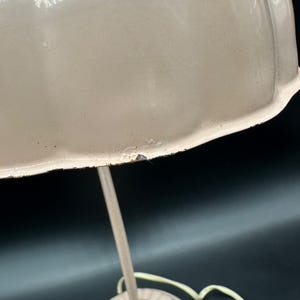 K&ouml;nnte beinhalten: Nahaufnahme einer cremefarbenen Vintage-Tischlampe. Die Lampe hat einen abgerundeten Schirm und einen schlanken, gebogenen Stiel. Der Schirm hat ein leicht abgenutztes Aussehen mit kleinen Unvollkommenheiten. Die Lampe steht vor einem dunklen Hintergrund.