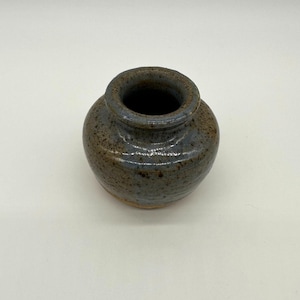 May include: A small, round ceramic vase with a wide, circular opening. The exterior features a mottled glaze with a blend of earthy brown and muted blue tones, characterized by a speckled texture throughout the surface of the clay vessel.