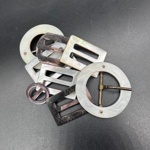 May include: A selection of vintage belt buckles in diverse shapes, including circular and rectangular. The buckles are crafted from iridescent mother-of-pearl and feature metal fittings. The colour scheme includes white, grey, and black.