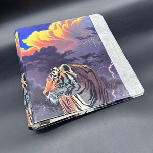 Puede incluir: Un archivador con una ilustración de un tigre sobre un cielo dramático con relámpagos y nubes. El tigre está representado en tonos marrones, naranjas y negros. El fondo presenta una vibrante puesta de sol y oscuras nubes de tormenta.