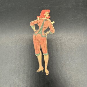 Puede incluir: Recorte de mu&ntilde;eca de papel vintage de una mujer con un atuendo rojo con ribetes verdes. La mu&ntilde;eca tiene el pelo rojo y posa con las manos en las caderas. El atuendo incluye una chaqueta y pantalones capri. La mu&ntilde;eca est&aacute; sobre un fondo negro.