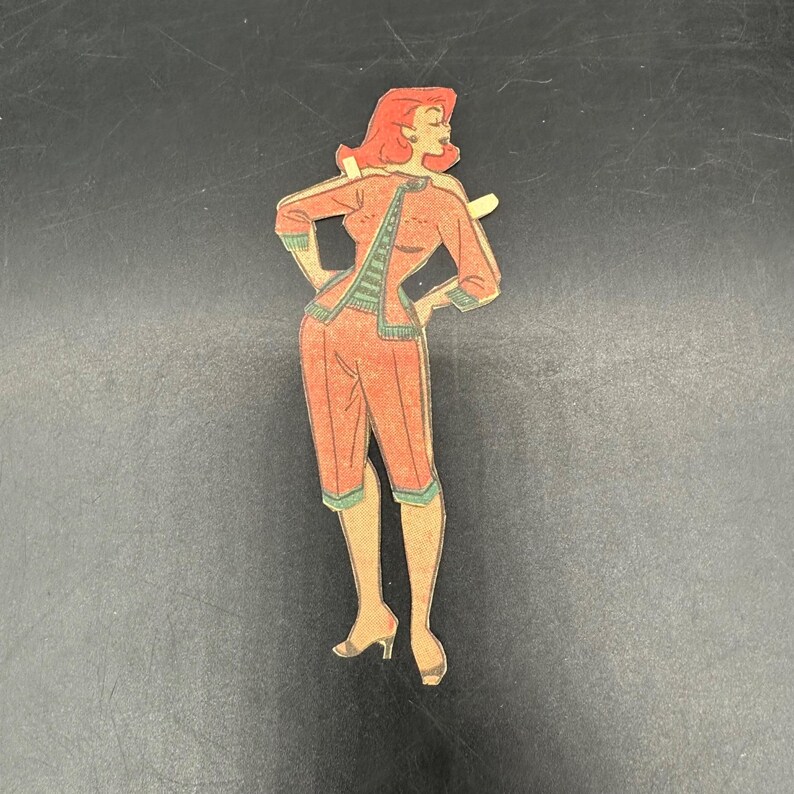 Puede incluir: Recorte de mu&ntilde;eca de papel vintage de una mujer con un atuendo rojo con ribetes verdes. La mu&ntilde;eca tiene el pelo rojo y posa con las manos en las caderas. El atuendo incluye una chaqueta y pantalones capri. La mu&ntilde;eca est&aacute; sobre un fondo negro.